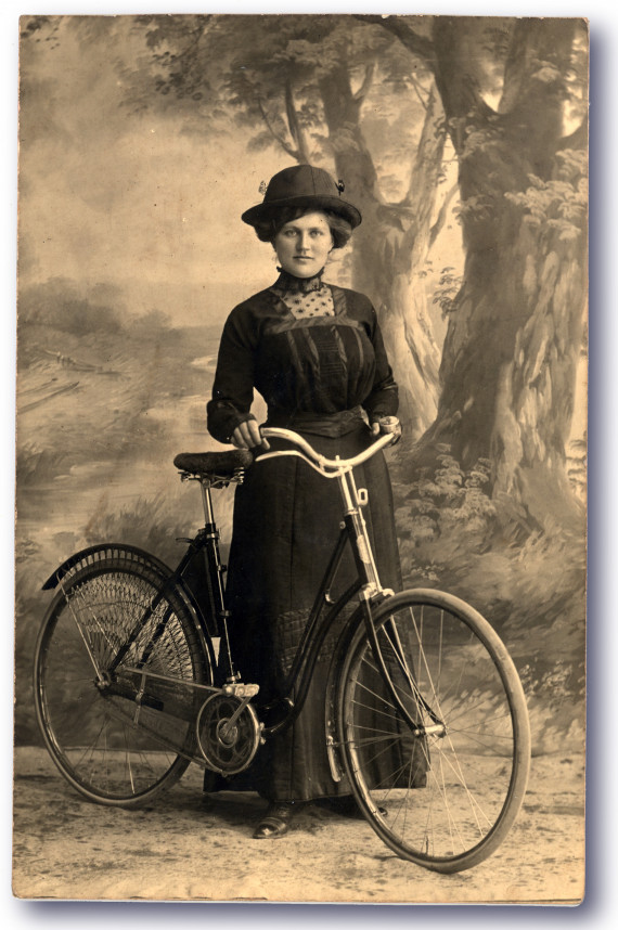 Vor einer auf eine Leinwand gemalten Landschaft mit Bäumen und Büschen steht eine Dame mit langem Rock und Hut hinter ihrem Fahrrad